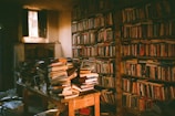 A dimly lit library room with tall bookshelves filled with numerous books. A small, rustic window allows a shaft of light to illuminate the room. In the center, a wooden table is piled high with stacks of books, some neatly arranged while others are in disarray. The ambiance is quiet and intimate, evoking a sense of history and knowledge.