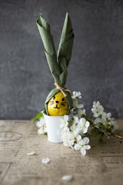 A yellow egg with a cute face is wrapped with a green napkin designed to resemble bunny ears, placed in a white pot. The arrangement is accompanied by white cherry blossoms on a surface that appears to be aged paper with text.