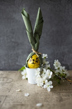 A yellow egg with a cute face is wrapped with a green napkin designed to resemble bunny ears, placed in a white pot. The arrangement is accompanied by white cherry blossoms on a surface that appears to be aged paper with text.