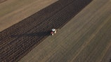 Aerial view of a tractor plowing a large agricultural field. The field is divided into distinct strips with the tractor working on a darker plowed section. The surrounding area is composed of lighter, untouched fields.