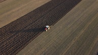 Aerial view of a tractor plowing a large agricultural field. The field is divided into distinct strips with the tractor working on a darker plowed section. The surrounding area is composed of lighter, untouched fields.