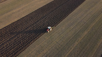 Aerial view of a tractor plowing a large agricultural field. The field is divided into distinct strips with the tractor working on a darker plowed section. The surrounding area is composed of lighter, untouched fields.