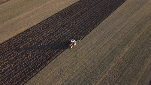 Aerial view of a tractor plowing a large agricultural field. The field is divided into distinct strips with the tractor working on a darker plowed section. The surrounding area is composed of lighter, untouched fields.