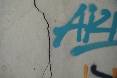 A textured concrete wall features a prominent vertical crack. On the right side of the image, there is graffiti composed of blue spray-painted lettering with a black and orange detail partially visible.