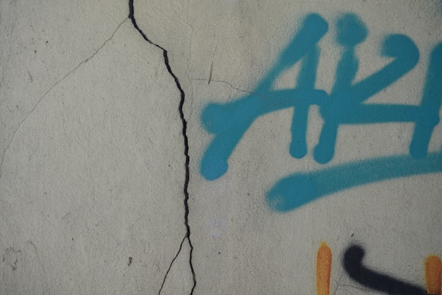 A textured concrete wall features a prominent vertical crack. On the right side of the image, there is graffiti composed of blue spray-painted lettering with a black and orange detail partially visible.