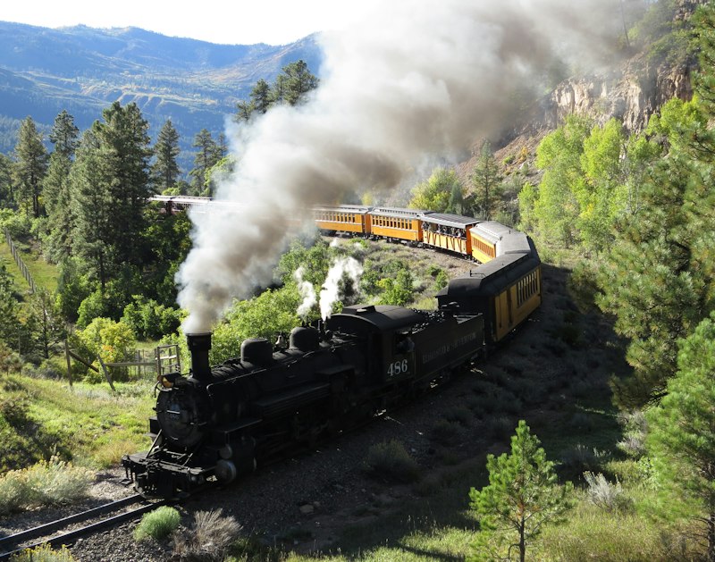 A black narrow-gauge steam locomotive pulling yellow coaches around a wooded bend with smoke billowing and mountains behind