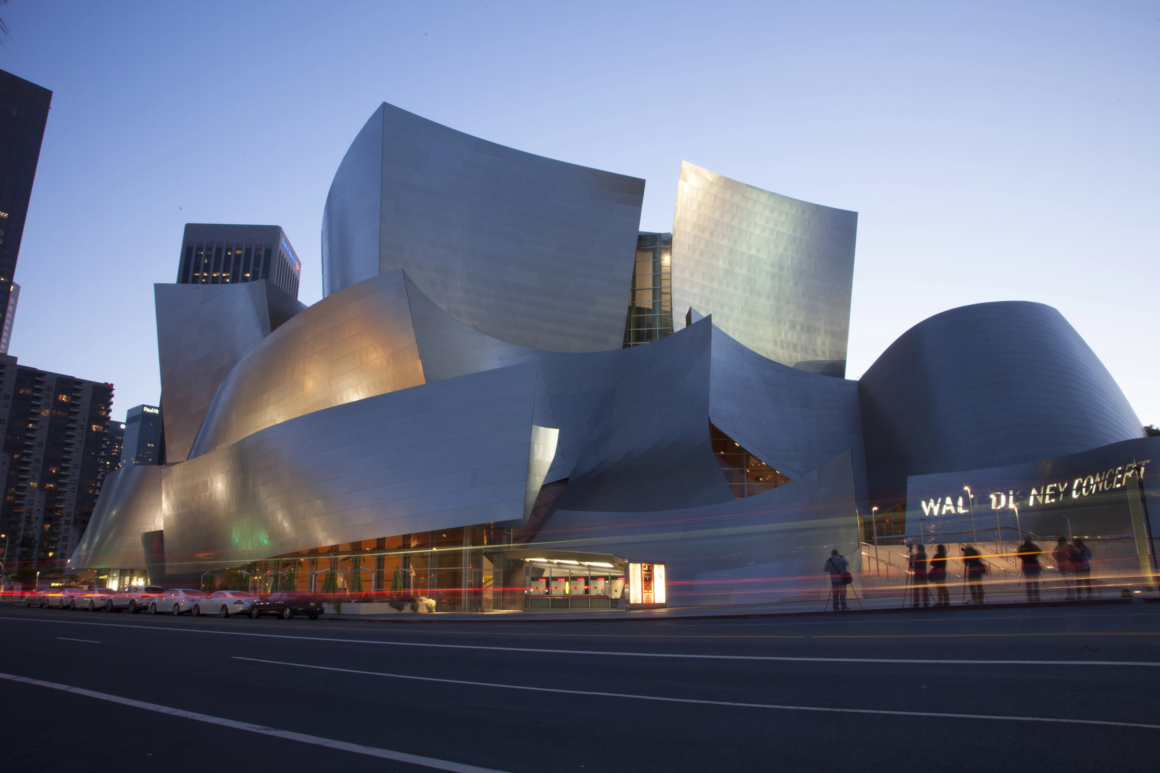 The billowing stainless-steel sails of the Walt Disney Concert Hall glowing silver and blue at dusk, its lit marquee and streaks of car light along Grand Avenue with downtown Los Angeles towers behind.