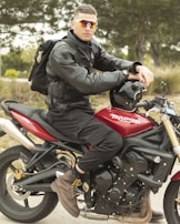 A person wearing a black leather jacket and sunglasses is sitting on a red Triumph motorcycle. The motorcycle is parked on a pathway surrounded by greenery. The individual is also wearing brown boots and has a black backpack.