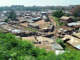 An aerial view of a densely populated informal settlement with rows of small, closely packed homes made of corrugated metal. The settlement is interspersed with narrow dirt pathways and some greenery, with trees and shrubs visible in the foreground and background. There are a few people walking along the pathways.