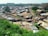 An aerial view of a densely populated informal settlement with rows of small, closely packed homes made of corrugated metal. The settlement is interspersed with narrow dirt pathways and some greenery, with trees and shrubs visible in the foreground and background. There are a few people walking along the pathways.