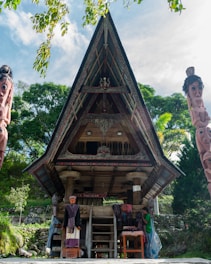 A traditional wooden structure with intricate carvings and a steeply angled roof dominates the scene, set against a backdrop of lush green trees. To the left and right, large wooden statues with carved faces are visible. At the base of the structure, a person is seen interacting with clothing items, possibly traditional fabrics, displayed on a table.