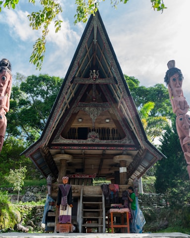 A traditional wooden structure with intricate carvings and a steeply angled roof dominates the scene, set against a backdrop of lush green trees. To the left and right, large wooden statues with carved faces are visible. At the base of the structure, a person is seen interacting with clothing items, possibly traditional fabrics, displayed on a table.