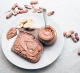 A white plate holds a slice of bread spread with creamy peanut butter. Nearby, there is a small jar of peanut butter with a spoon inside. Slices of banana are placed on the plate, adding a touch of freshness. Scattered around are whole and shelled peanuts, contributing to a rustic presentation.