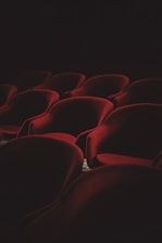 A dimly lit interior showcasing rows of plush, red velvet theater seats arranged in a pattern. The lighting casts soft shadows that enhance the luxurious texture of the fabric, contributing to an atmosphere of quiet anticipation.