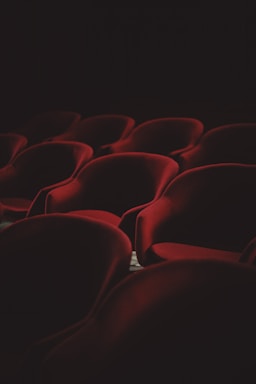 A dimly lit interior showcasing rows of plush, red velvet theater seats arranged in a pattern. The lighting casts soft shadows that enhance the luxurious texture of the fabric, contributing to an atmosphere of quiet anticipation.