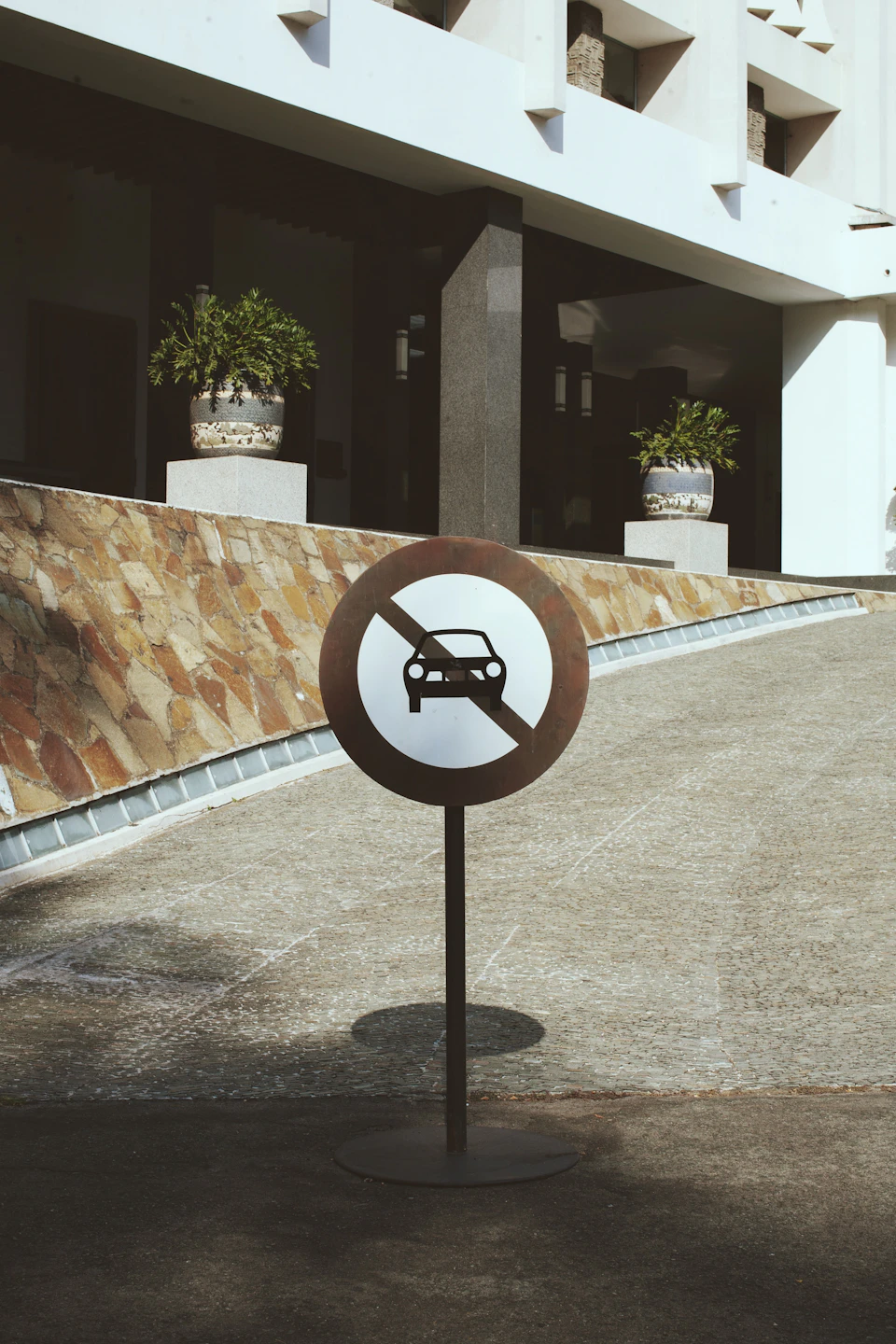 Circular sign with white border and black car silhouette mounted on metal pole against stone