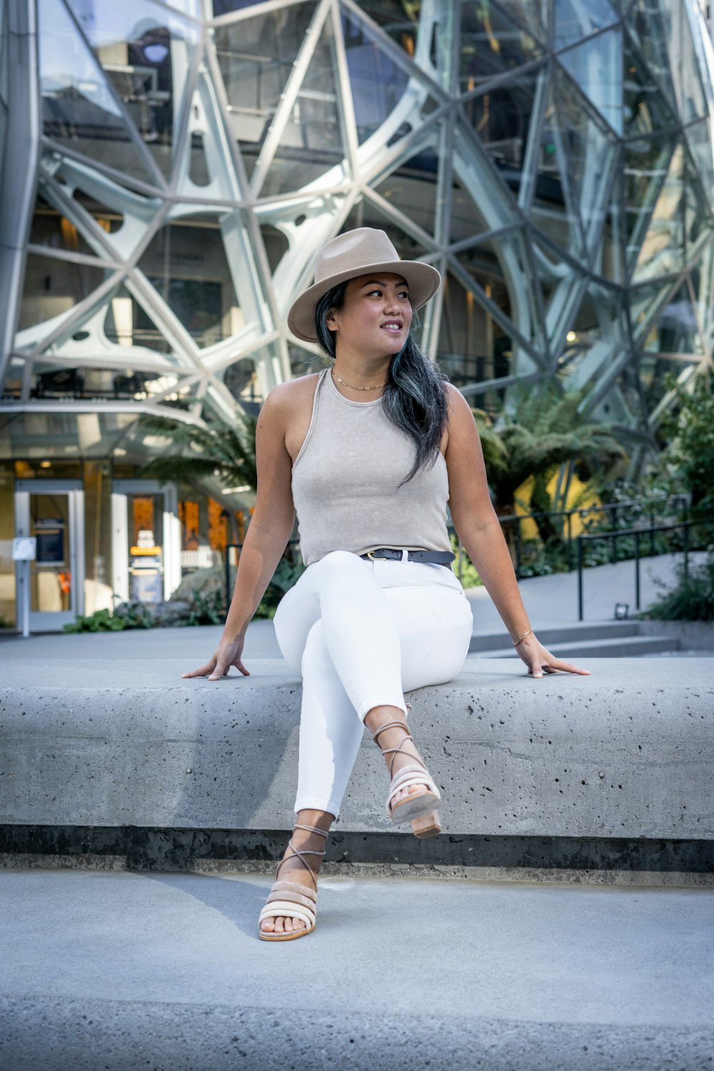 Mujer con camiseta blanca sin mangas y pantalones blancos con sombrero marrón sentada en un banco blanco