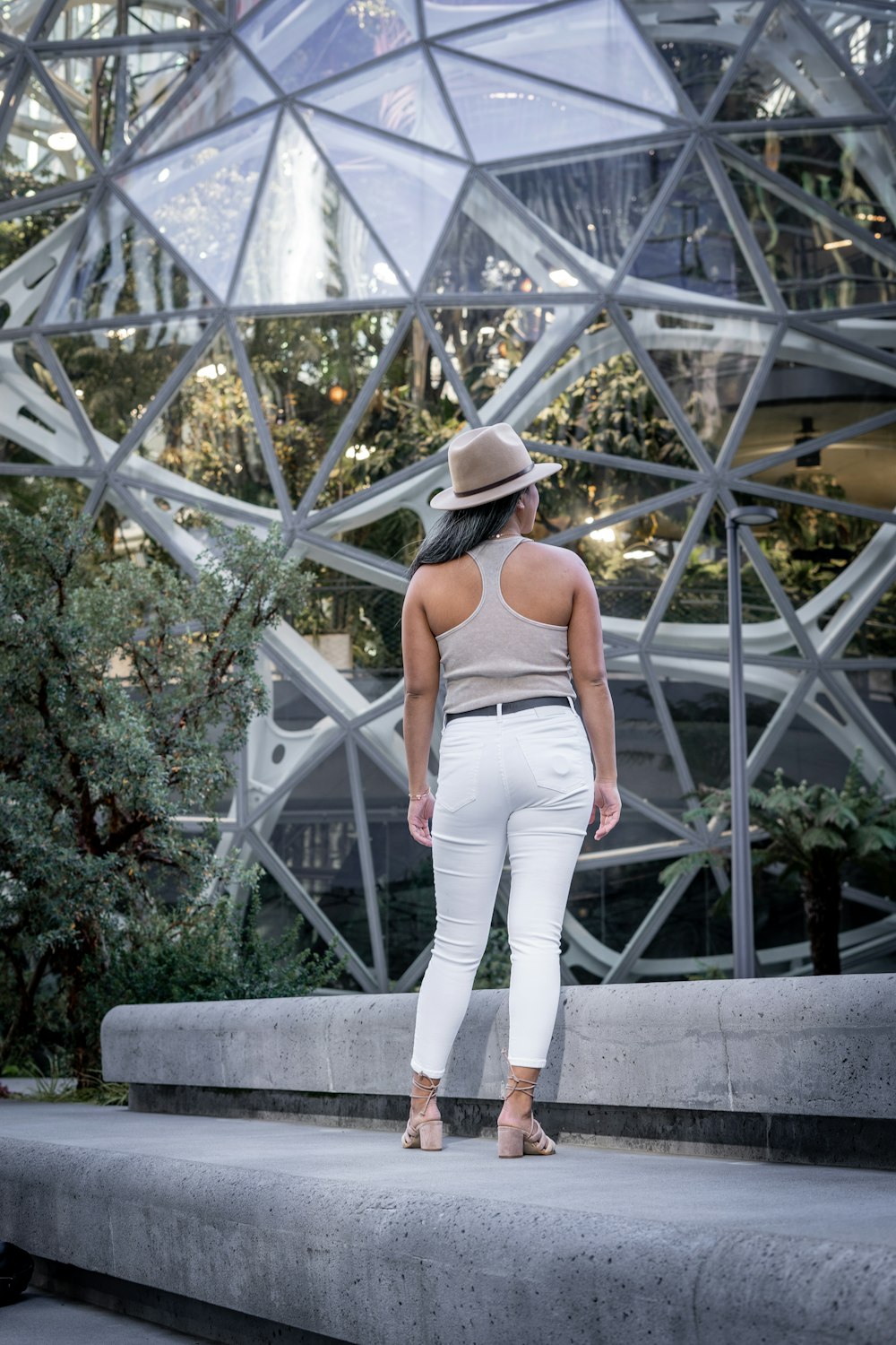 Femme en débardeur gris et pantalon blanc portant un chapeau fedora noir debout sur du béton gris