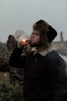 A person wearing a dark jacket with a fur-lined hood stands outdoors, holding and lighting a cigar. The background is hazy with a log or large piece of wood visible.