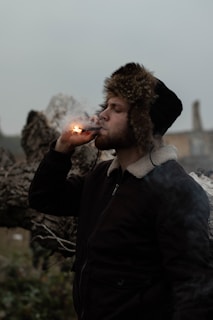 A person wearing a dark jacket with a fur-lined hood stands outdoors, holding and lighting a cigar. The background is hazy with a log or large piece of wood visible.
