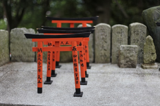 A series of small red torii gates with black tops are lined up on a stone surface, surrounded by stone tablets with vertical inscriptions. The background is filled with greenery, creating a serene and spiritual atmosphere.