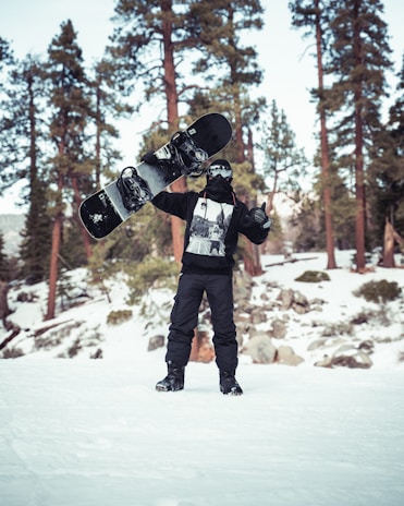 A person dressed in winter gear holding a snowboard vertically in a snowy wooded area. The person is wearing a hoodie and ski mask, surrounded by snow-covered ground and tall evergreen trees.