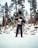 A person dressed in winter gear holding a snowboard vertically in a snowy wooded area. The person is wearing a hoodie and ski mask, surrounded by snow-covered ground and tall evergreen trees.