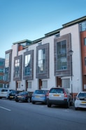 Modern urban residential building featuring large glass windows and contemporary architecture. Several parked cars are lined up on the street in front of the building, which has clean, white walls and wooden accents around the windows.