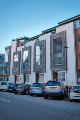 Modern urban residential building featuring large glass windows and contemporary architecture. Several parked cars are lined up on the street in front of the building, which has clean, white walls and wooden accents around the windows.