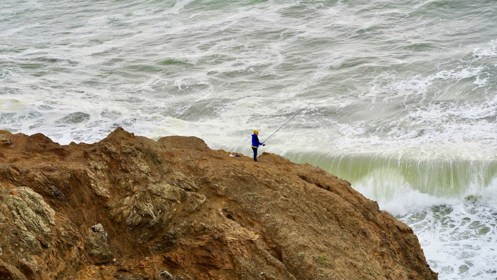 man in blue shirt and blue pants standing on brown rock formation near body of water