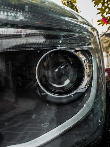Close-up view of a car headlight with reflective surfaces and smooth curves. The intricate details of the glass and surrounding metal are visible, with some greenery and a pink flower blurred in the background.