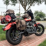 A motorcycle is parked on a brick pavement, heavily loaded with various brown and black bags and a rolled-up red, white, and black blanket. The background features green trees, a grassy lawn, some residential houses, and utility poles with power lines.