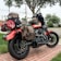 A motorcycle is parked on a brick pavement, heavily loaded with various brown and black bags and a rolled-up red, white, and black blanket. The background features green trees, a grassy lawn, some residential houses, and utility poles with power lines.