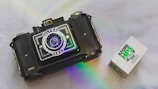 A vintage camera with a prominent lens and detailed body design is positioned on a smooth surface. Beside it is a box of Ilford HP5 Plus 400 black and white film. A subtle rainbow reflection adds a touch of color to the scene.