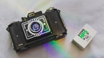 A vintage camera with a prominent lens and detailed body design is positioned on a smooth surface. Beside it is a box of Ilford HP5 Plus 400 black and white film. A subtle rainbow reflection adds a touch of color to the scene.
