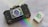 A vintage camera with a prominent lens and detailed body design is positioned on a smooth surface. Beside it is a box of Ilford HP5 Plus 400 black and white film. A subtle rainbow reflection adds a touch of color to the scene.