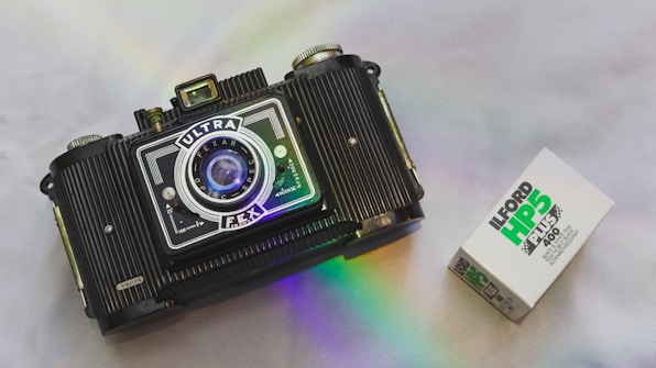 A vintage camera with a prominent lens and detailed body design is positioned on a smooth surface. Beside it is a box of Ilford HP5 Plus 400 black and white film. A subtle rainbow reflection adds a touch of color to the scene.