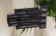 A stack of black cosmetic product boxes labeled with 'HD Brows' and various product names such as Brow Colourfix, Browtec, and Brow Define. The boxes are reflected on a glass-like surface, with a blurred green plant in the background.