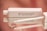 A white rectangular cosmetic tube with the brand name 'HD Brows' and the text 'High Definition Beauty' printed on it lies on a reflective surface, its cap removed and a white applicator wand placed beside it. Water droplets are visible on the surface, adding a glossy appearance. The background features a soft pink gradient.