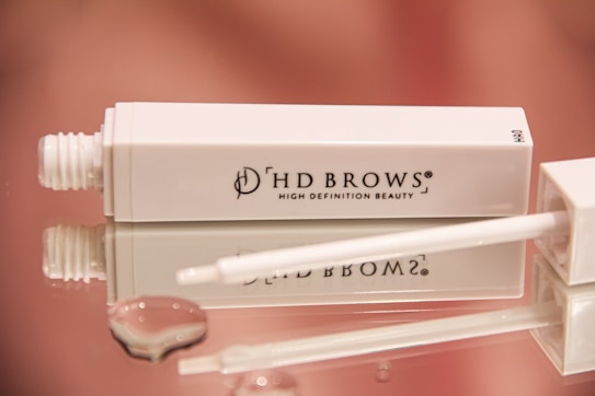 A white rectangular cosmetic tube with the brand name 'HD Brows' and the text 'High Definition Beauty' printed on it lies on a reflective surface, its cap removed and a white applicator wand placed beside it. Water droplets are visible on the surface, adding a glossy appearance. The background features a soft pink gradient.