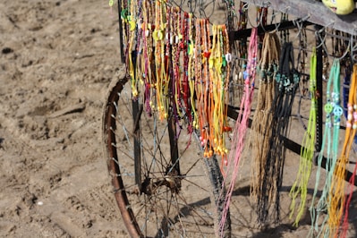 Colorful handmade necklaces and bracelets hang from a metal frame next to a sandy beach. The frame is part of an old bicycle wheel, and the strands vary in color, including vibrant yellows, oranges, pinks, and blues.