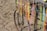Colorful handmade necklaces and bracelets hang from a metal frame next to a sandy beach. The frame is part of an old bicycle wheel, and the strands vary in color, including vibrant yellows, oranges, pinks, and blues.