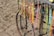 Colorful handmade necklaces and bracelets hang from a metal frame next to a sandy beach. The frame is part of an old bicycle wheel, and the strands vary in color, including vibrant yellows, oranges, pinks, and blues.