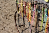 Colorful handmade necklaces and bracelets hang from a metal frame next to a sandy beach. The frame is part of an old bicycle wheel, and the strands vary in color, including vibrant yellows, oranges, pinks, and blues.