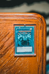 A collectible card encased in a protective sleeve is displayed on a wooden surface. The card features a blue and white illustration of a powerful creature labeled 'Obelisk the Tormentor'. The card text includes details about the creature's attributes such as attack and defense points.