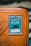 A collectible card encased in a protective sleeve is displayed on a wooden surface. The card features a blue and white illustration of a powerful creature labeled 'Obelisk the Tormentor'. The card text includes details about the creature's attributes such as attack and defense points.