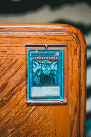 A collectible card encased in a protective sleeve is displayed on a wooden surface. The card features a blue and white illustration of a powerful creature labeled 'Obelisk the Tormentor'. The card text includes details about the creature's attributes such as attack and defense points.