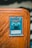 A collectible card encased in a protective sleeve is displayed on a wooden surface. The card features a blue and white illustration of a powerful creature labeled 'Obelisk the Tormentor'. The card text includes details about the creature's attributes such as attack and defense points.