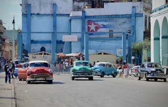 A vibrant street scene with classic cars parked in front of a weathered, blue-painted building with a mural of a flag. People are walking along the sidewalk, and the atmosphere feels lively and bustling.