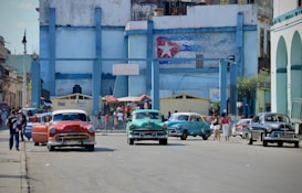 A vibrant street scene with classic cars parked in front of a weathered, blue-painted building with a mural of a flag. People are walking along the sidewalk, and the atmosphere feels lively and bustling.